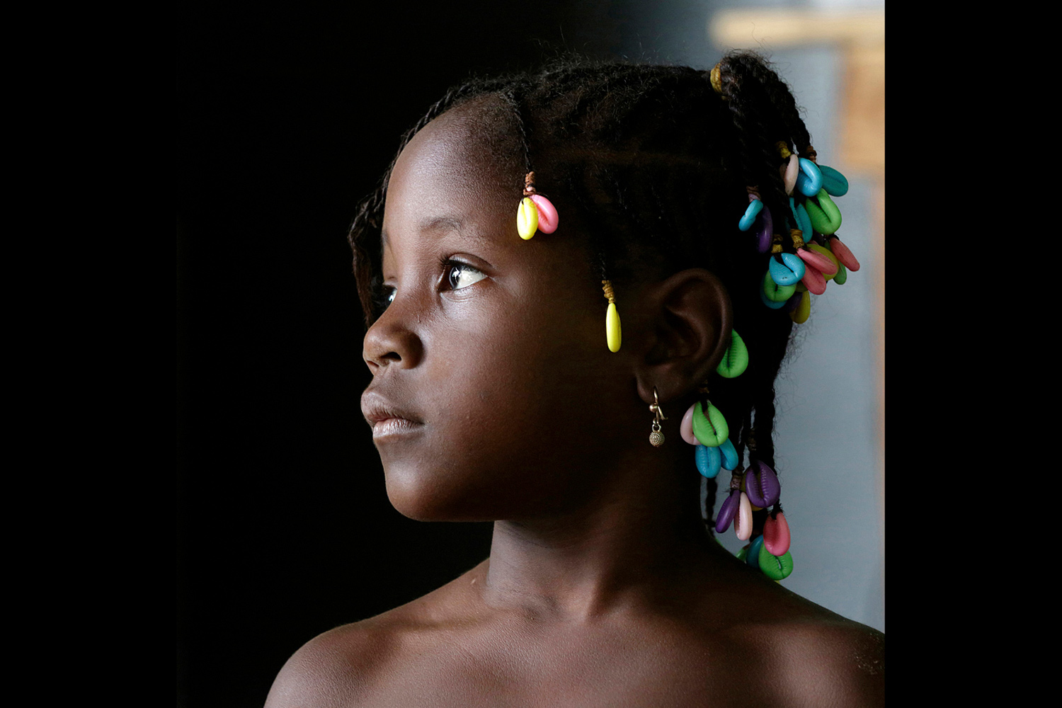 Jeune fille au Bénin en 2015 avec des cauris colorés dans les cheveux, regard sur le côté