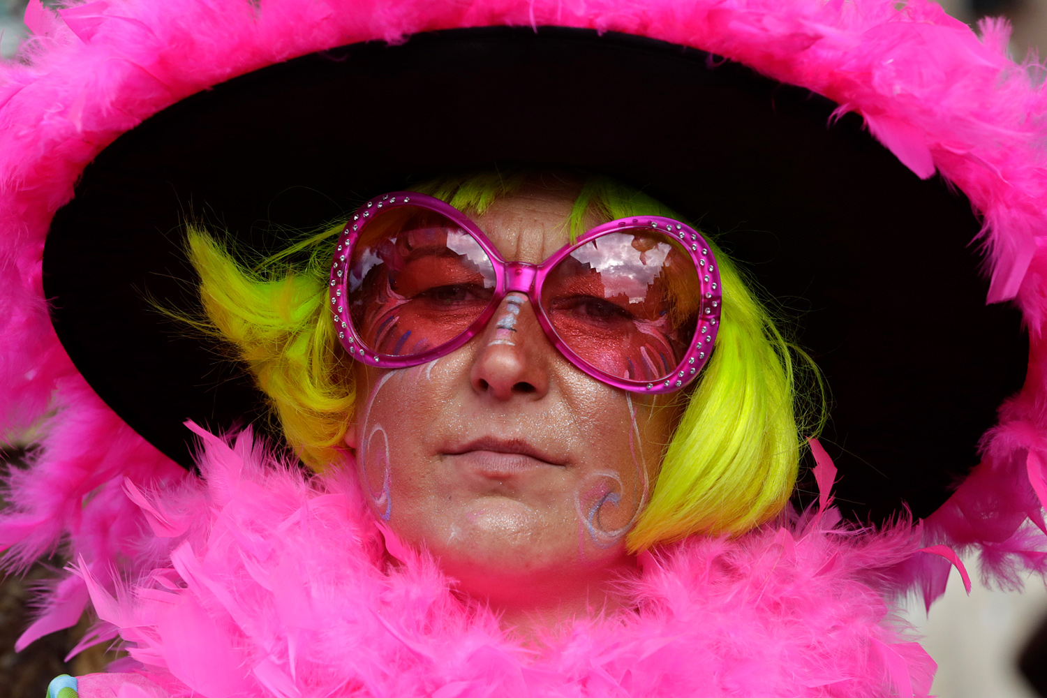 Portrait d'une carnavalière portant une grande capeline noire bordée de plumes roses, une perruque jaune et des lunettes roses oversize.