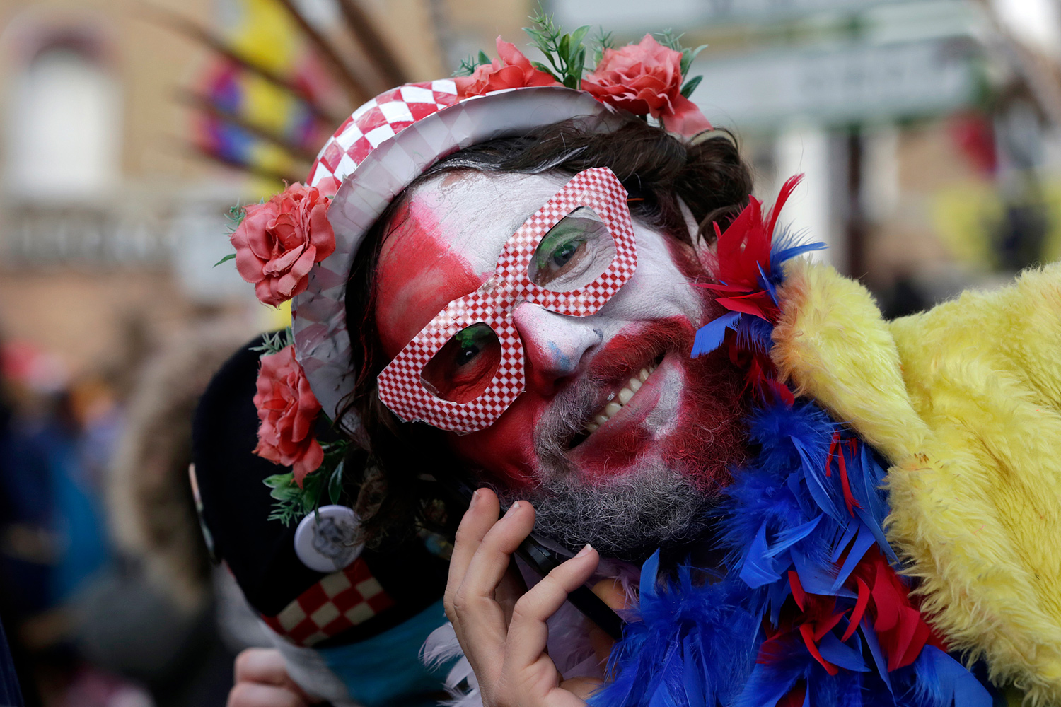 Portrait d'un carnavaleux maquillé de rouge, blanc et noir, portant un masque en résille, un chapeau décoré de fleurs et un boa jaune.