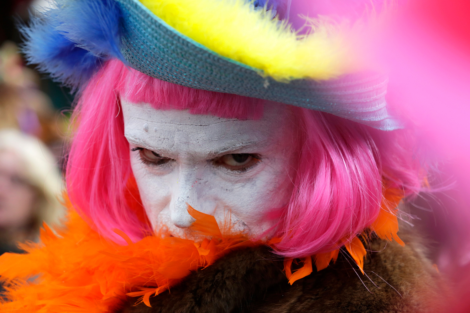 Portrait d'un carnavaleux au visage maquillé de blanc, portant une perruque rose, un chapeau bleu et jaune et un boa orange.