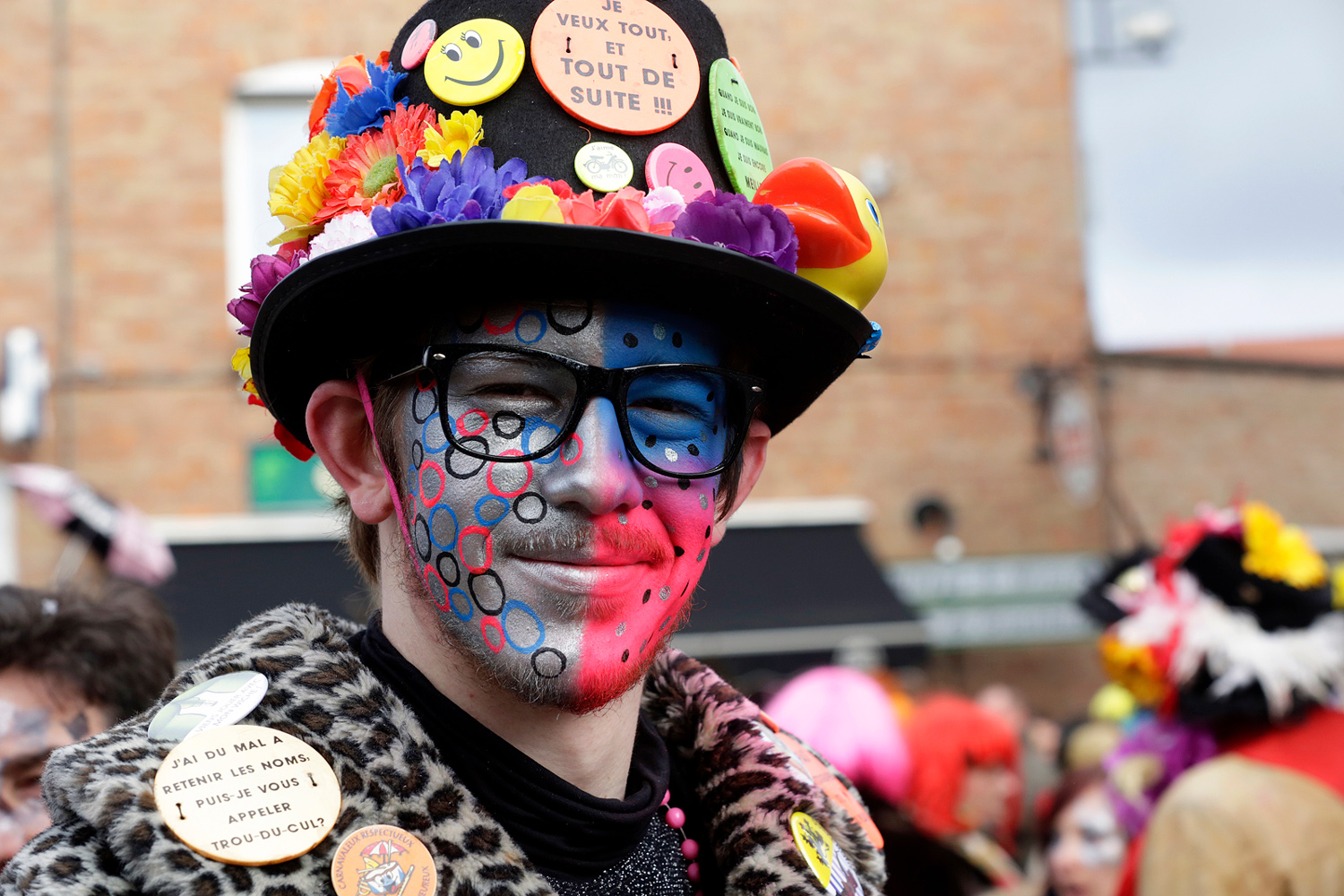 Portrait d'un carnavaleux portant un chapeau décoré de nombreux badges et un maquillage bleu, rouge et noir aux motifs circulaires.