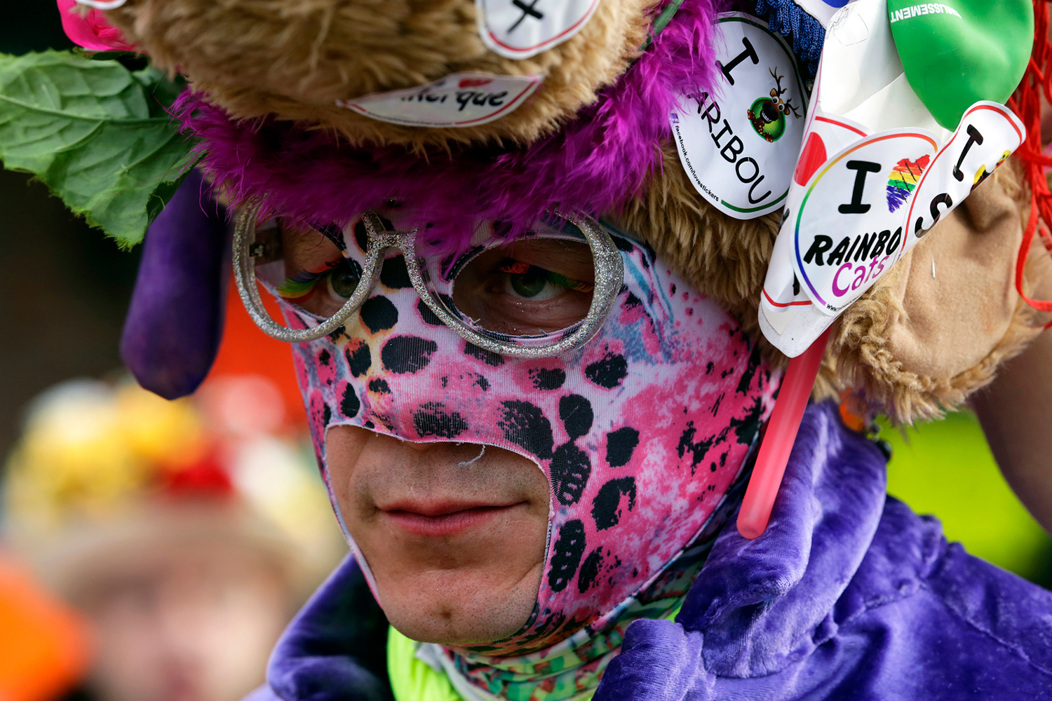 Portrait d'un carnavaleux portant un chapeau décoré de peluches, un maquillage rose et noir à motifs tachetés et un costume violet.