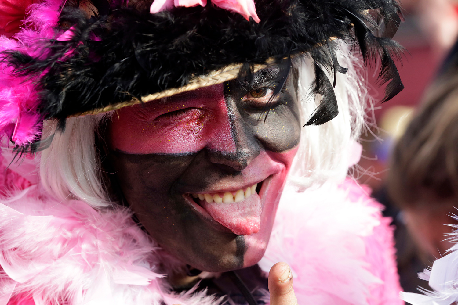 Portrait d'un carnavaleux maquillé de noir et de rose, coiffé d'un chapeau orné de plumes noires et roses, tirant la langue.