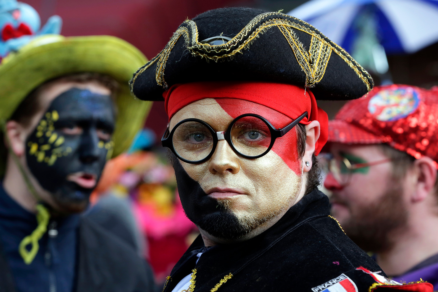 Portrait d'un carnavaleux portant un tricorne noir, des lunettes rondes et un maquillage blanc, rouge et noir.