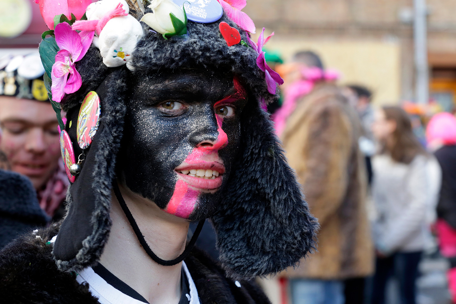 Portrait d'une carnavaleux maquillé de noir et de rose, portant une chapka noire décorée de fleurs et de badges.