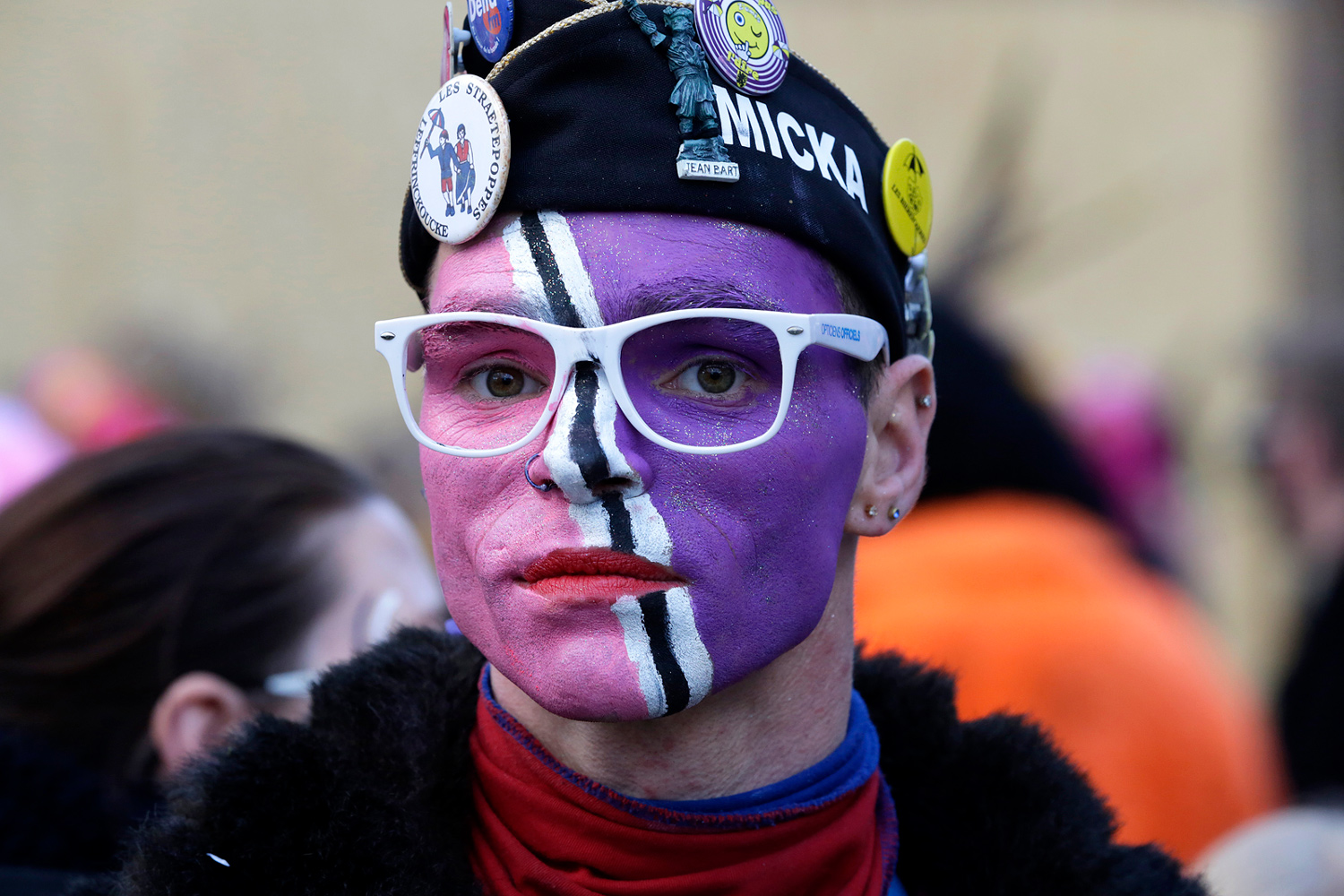Portrait d'un carnavaleux portant un béret noir décoré de badges, des lunettes blanches et un maquillage violet et blanc.