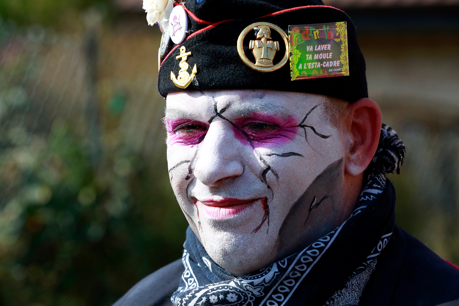 Portrait d'un carnavaleux maquillé de blanc avec des traits noirs et violets, coiffé d'un béret noir orné d'insignes et d'un foulard noir.