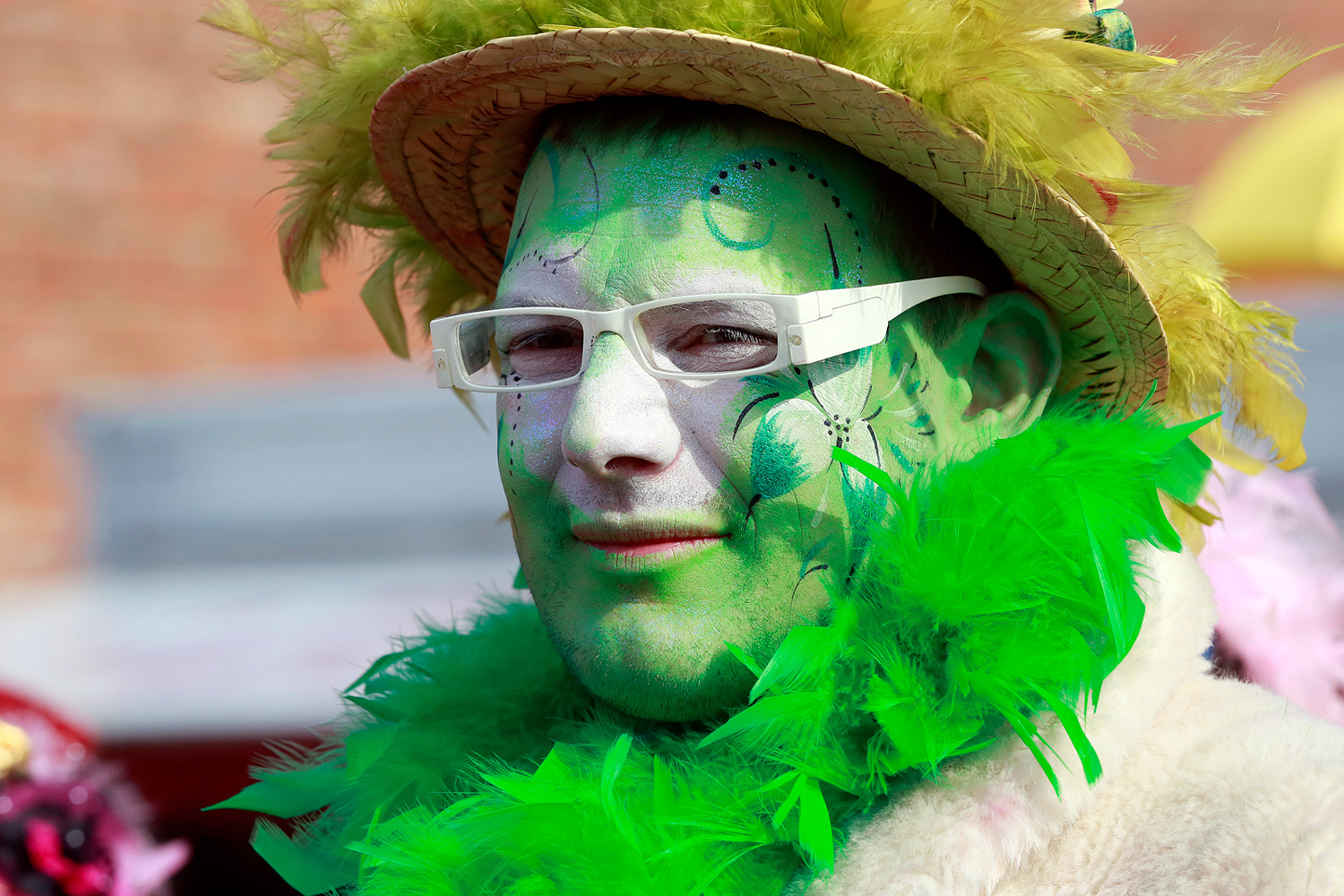 Portrait d'un carnavaleux portant un chapeau de paille, des lunettes blanches, un maquillage vert et blanc et un boa de plumes vertes, lors de la Bande de Pitgam du Carnaval de Dunkerque.