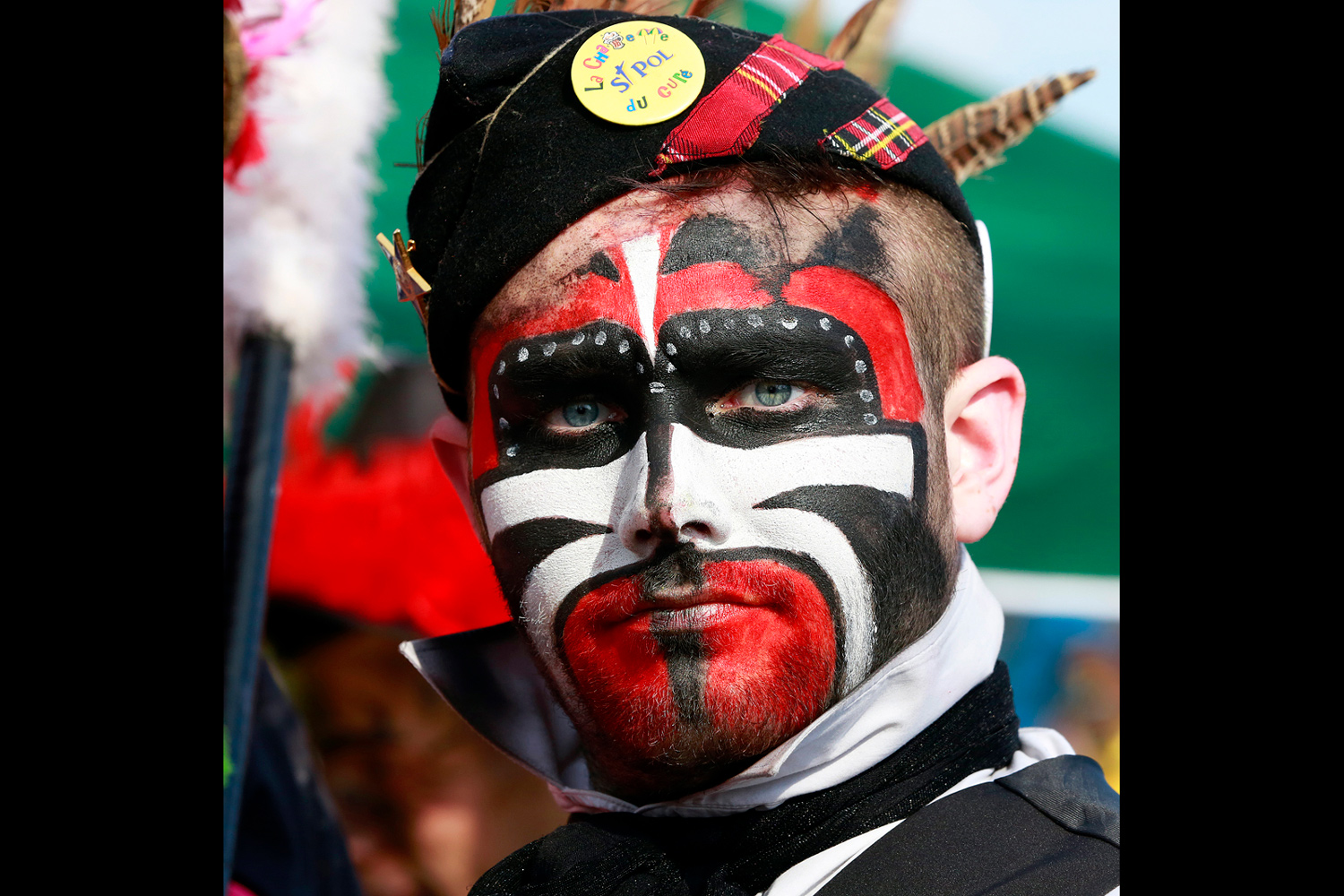 Portrait d'un carnavaleux portant un béret noir décoré de badges et un maquillage rouge, noir et blanc aux formes géométriques.