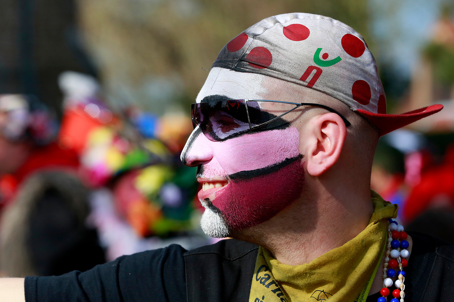 Portrait de profil d'un carnavaleux portant une casquette blanche à pois rouges, des lunettes noires et un maquillage rose et blanc.