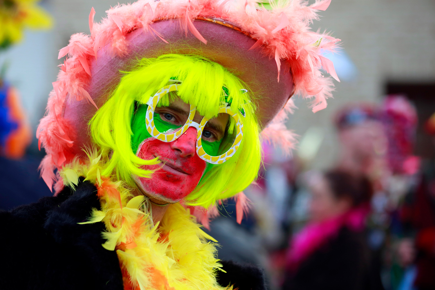 Portrait d'un carnavaleux portant une perruque jaune fluo, un maquillage rouge, blanc et vert, des lunettes fantaisie vertes et un chapeau rose orné de plumes, lors de la Bande de Pitgam du Carnaval de Dunkerque.