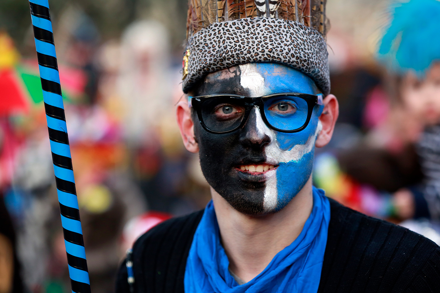 Portrait d'un carnavaleux portant un maquillage bleu, noir et blanc, coiffé d'une couronne tressée et tenant une canne rayée.