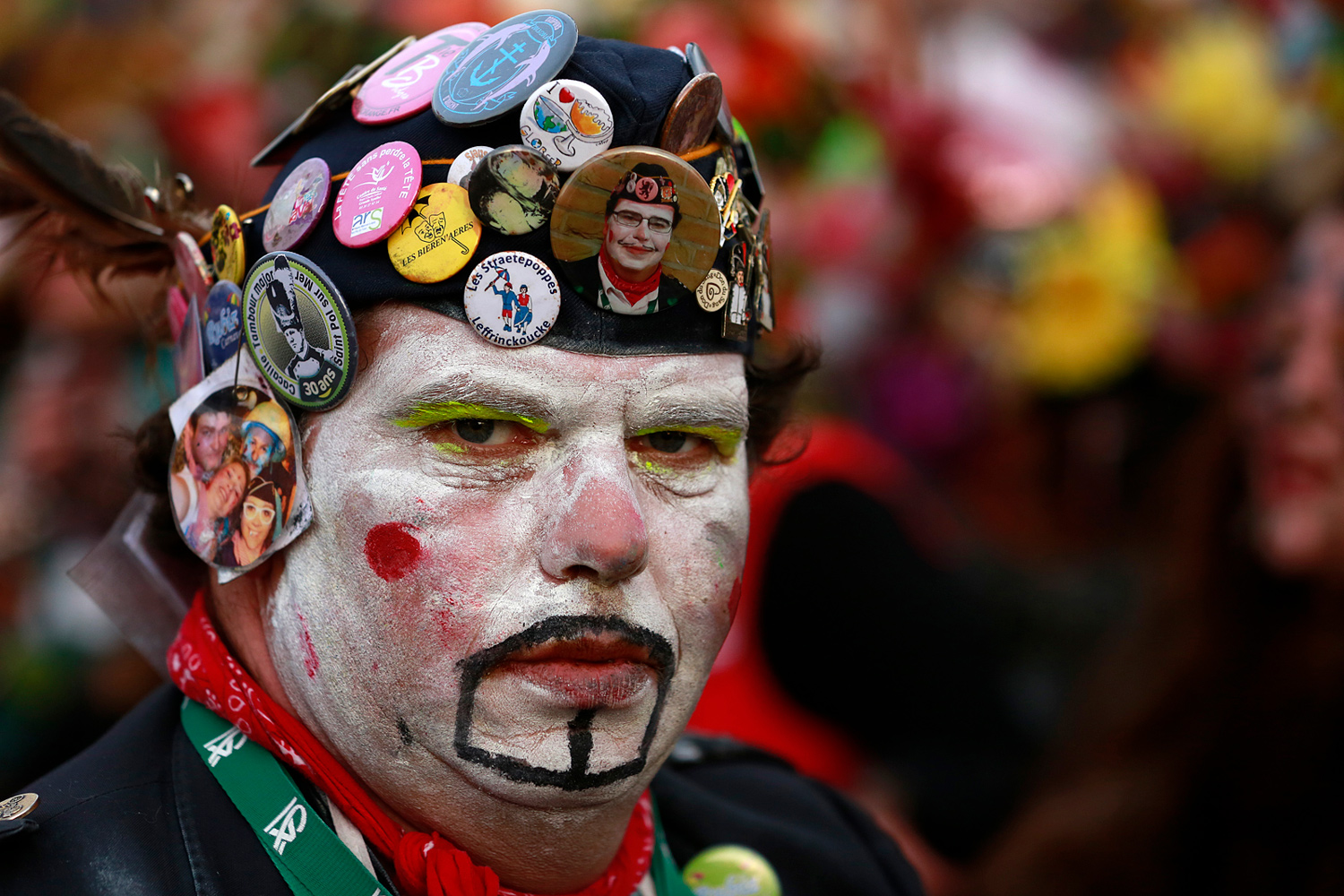 Portrait d'un carnavaleux au visage maquillé de blanc, coiffé d'un béret noir recouvert de nombreux badges colorés.