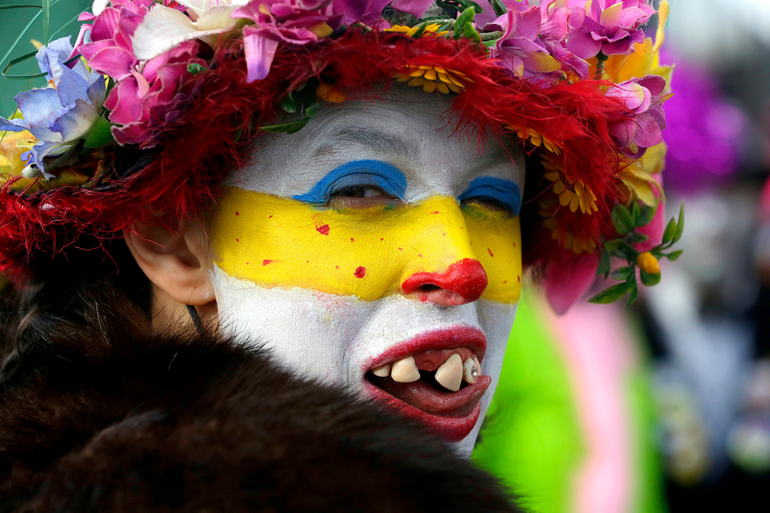 Portrait d'une carnavalière portant un maquillage blanc, jaune et bleu, un chapeau fleuri et des fausses dents, lors de la Bande de Bergues du Carnaval de Dunkerque.