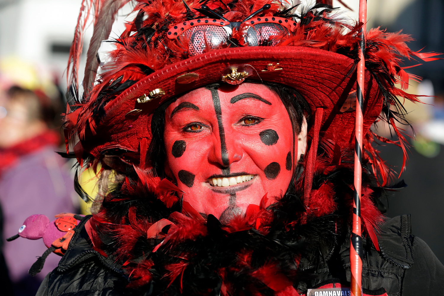 Portrait d'une carnavalière portant un large chapeau rouge décoré de plumes et un maquillage rouge orné de pois noirs.
