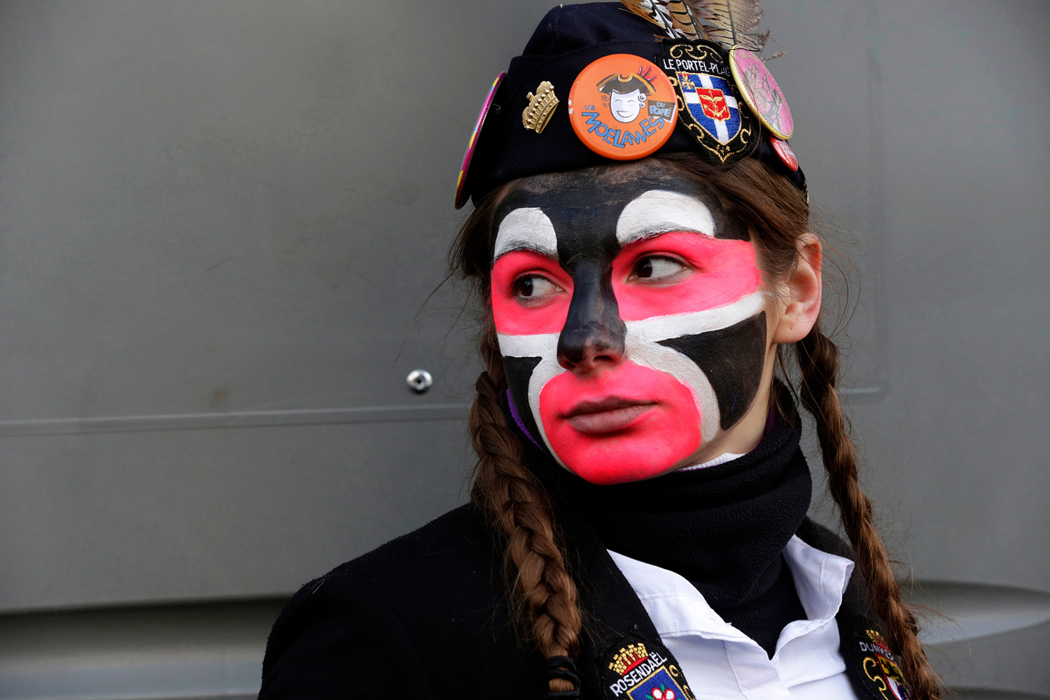 Portrait d'une carnavalière portant un béret noir décoré de badges et un maquillage rouge, noir et blanc, regardant vers la droite.