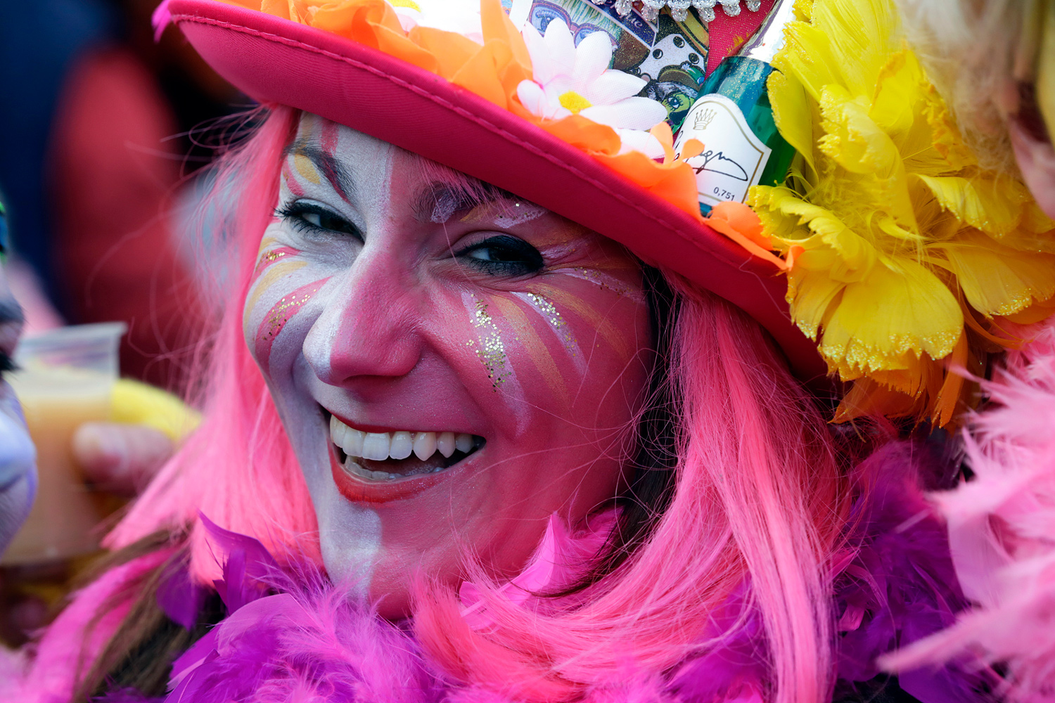 Portrait d'une carnavalière souriante portant une perruque rose, un chapeau orange fleuri et un maquillage rose pailleté.