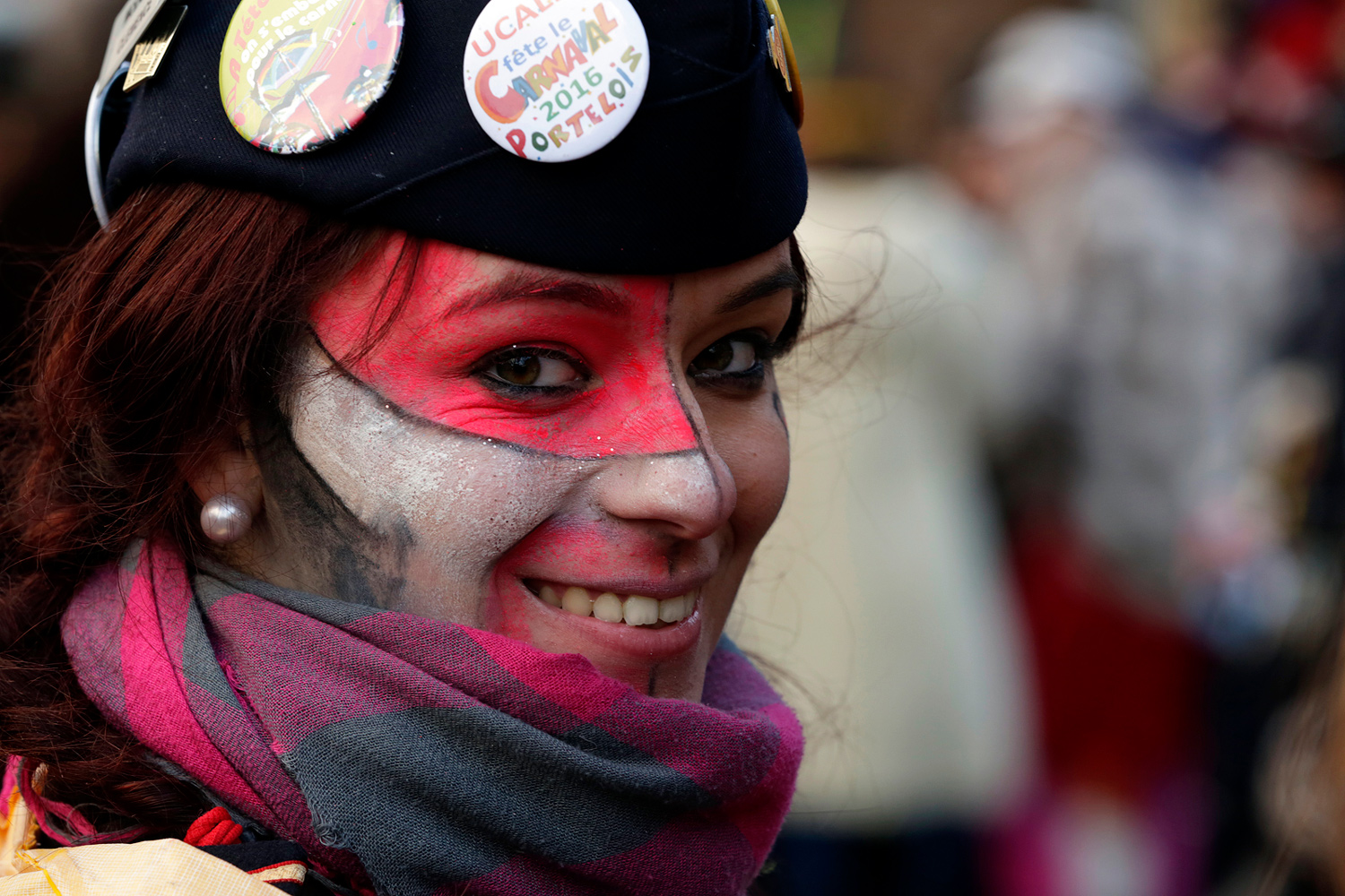 Portrait d'une carnavalière portant un béret noir décoré de badges et un maquillage rouge, noir et blanc.