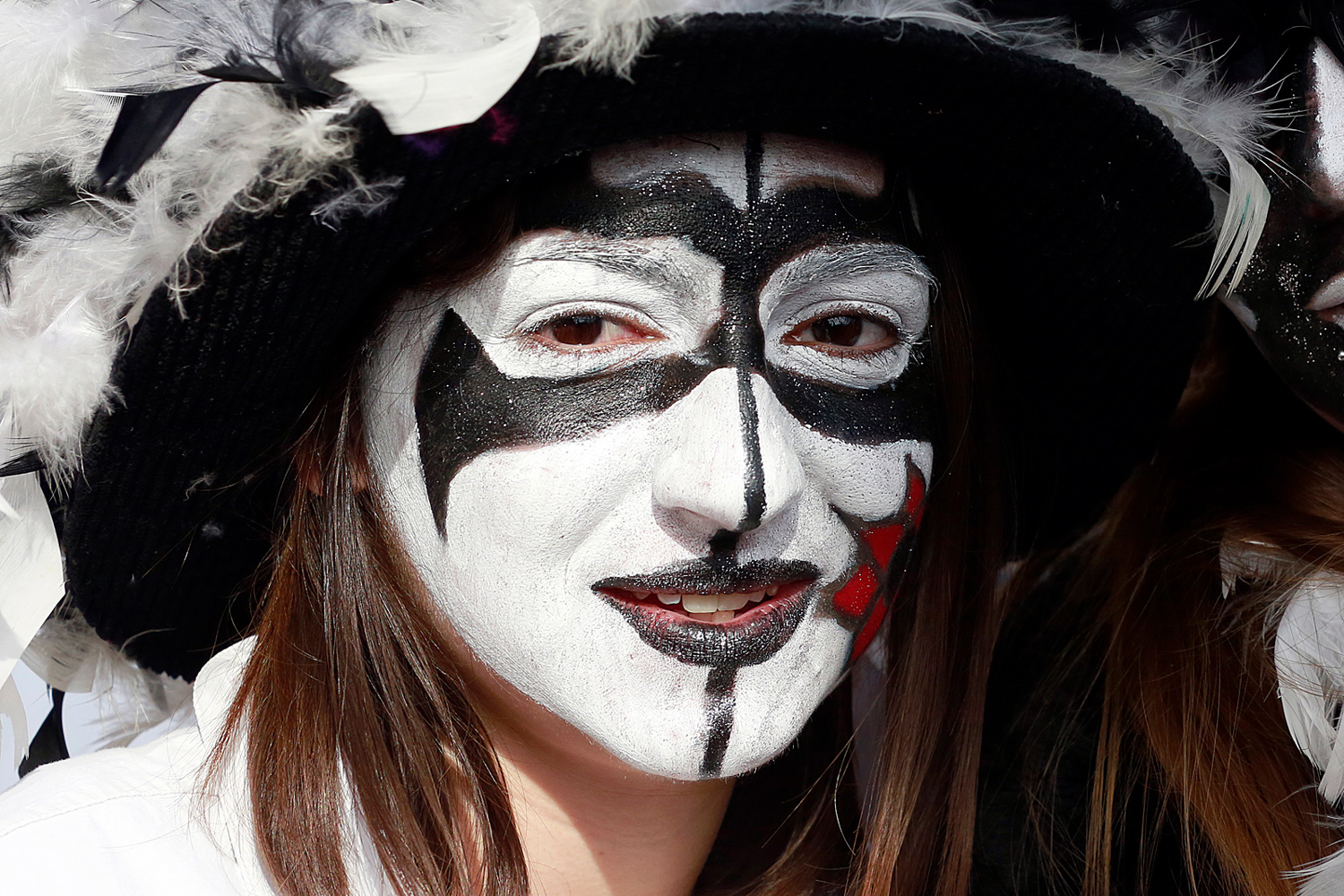 Portrait d'une carnavalière portant un maquillage noir et blanc aux motifs graphiques, coiffée d'un chapeau noir orné de plumes blanches, lors de la Bande de Pitgam du Carnaval de Dunkerque.