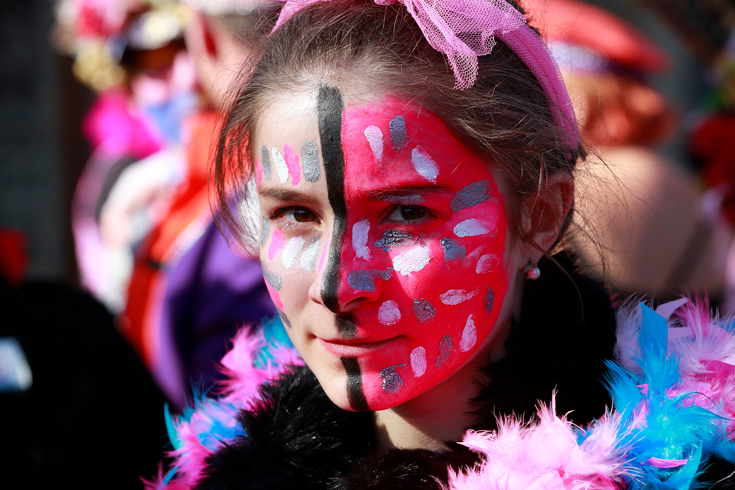 Portrait d'une carnavalière dont le visage est maquillé de rose, de blanc et de rouge, avec un serre-tête orné de plumes roses.