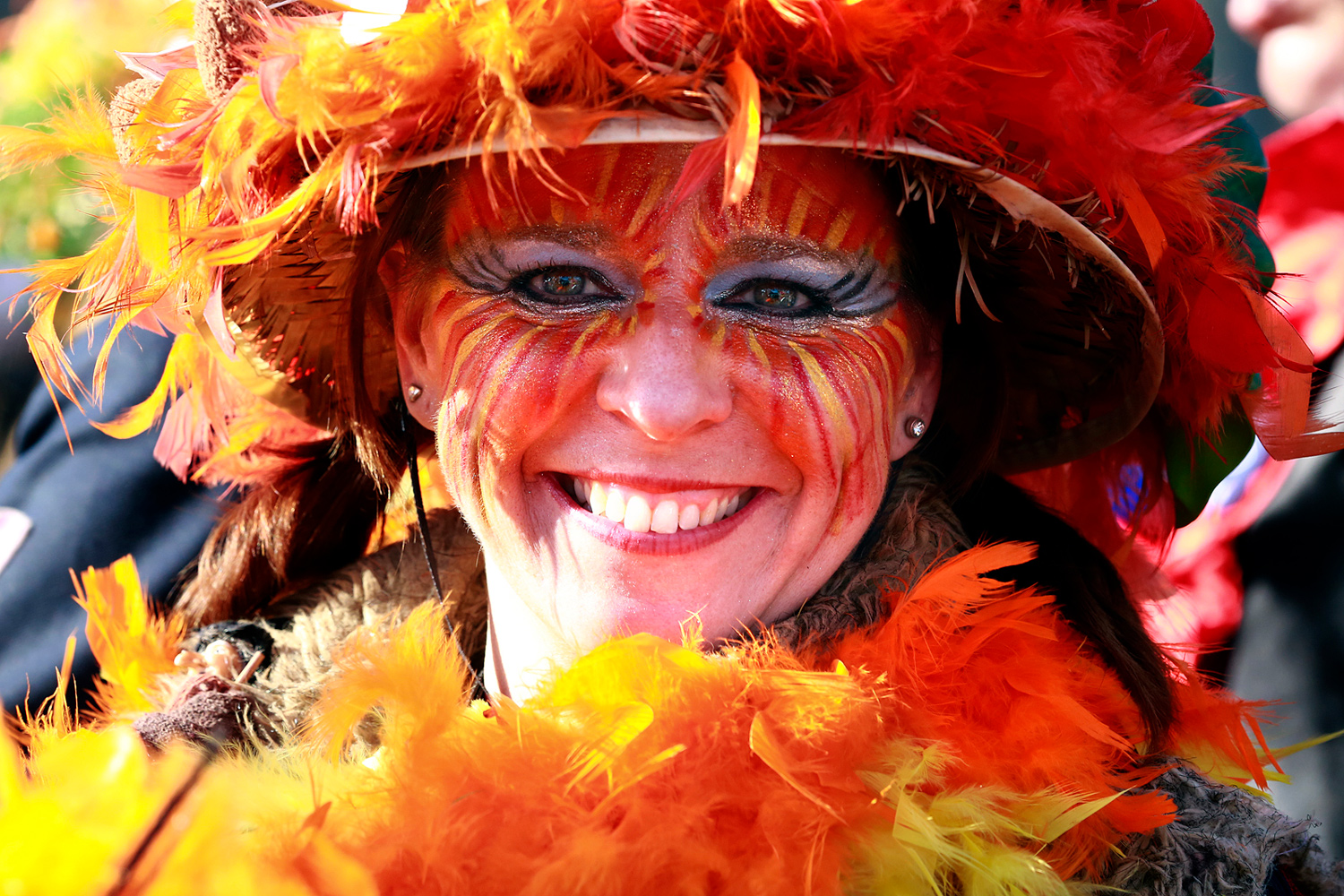 Portrait d'une carnavalière souriante portant un chapeau orange orné de plumes et un maquillage assorti aux couleurs chaudes.