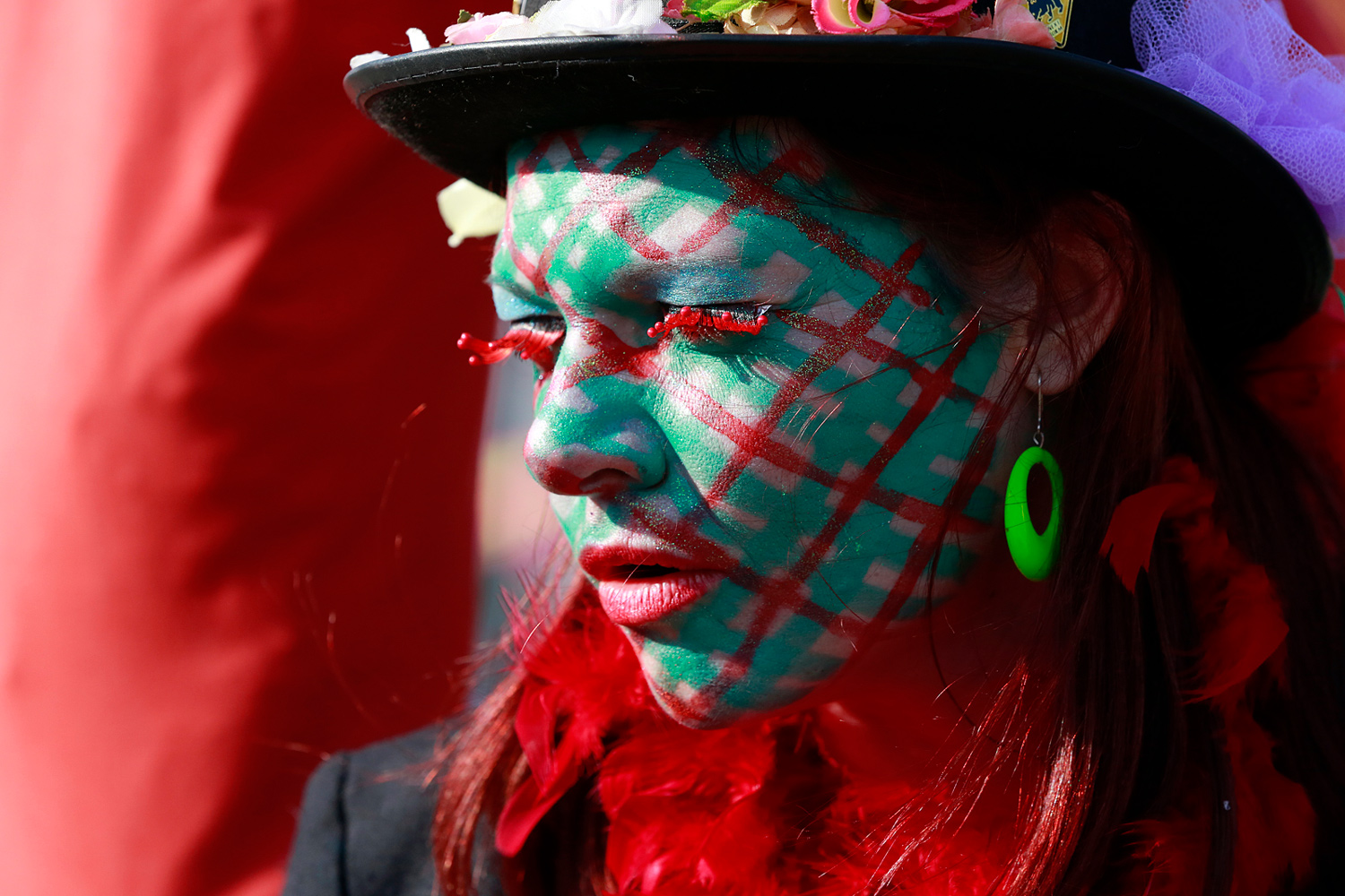 Portrait d'une carnavalière au visage maquillé de vert et de rouge avec un quadrillage rouge, portant un chapeau noir et une boucle d'oreille verte.