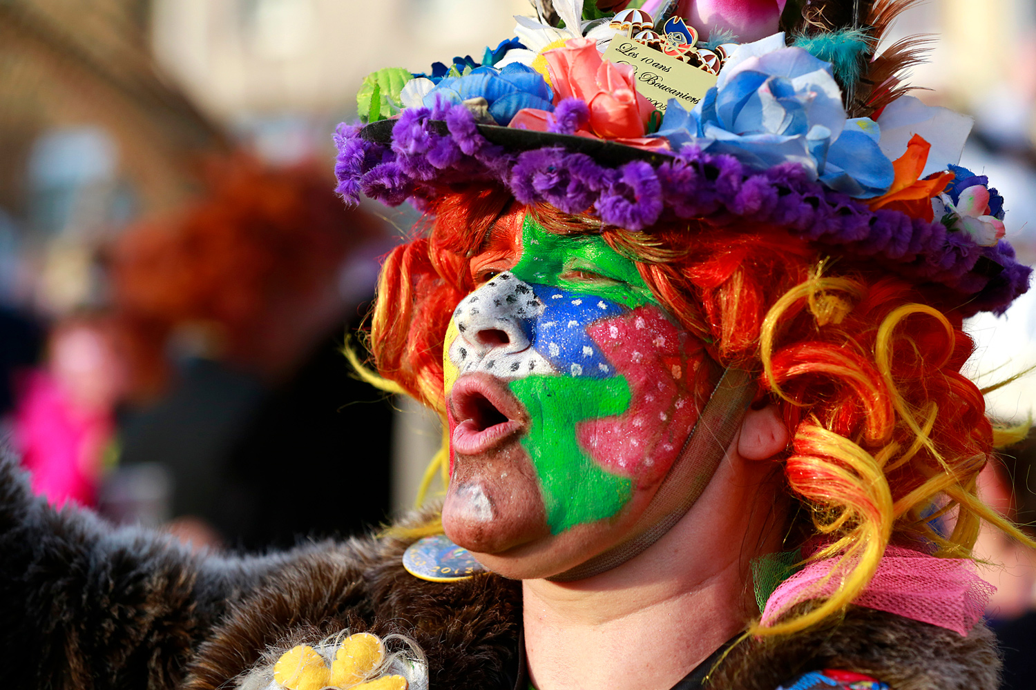 Portrait de profil d'une carnavalière au visage peint de vert, rouge et blanc, coiffée d'un chapeau fleuri aux couleurs vives.