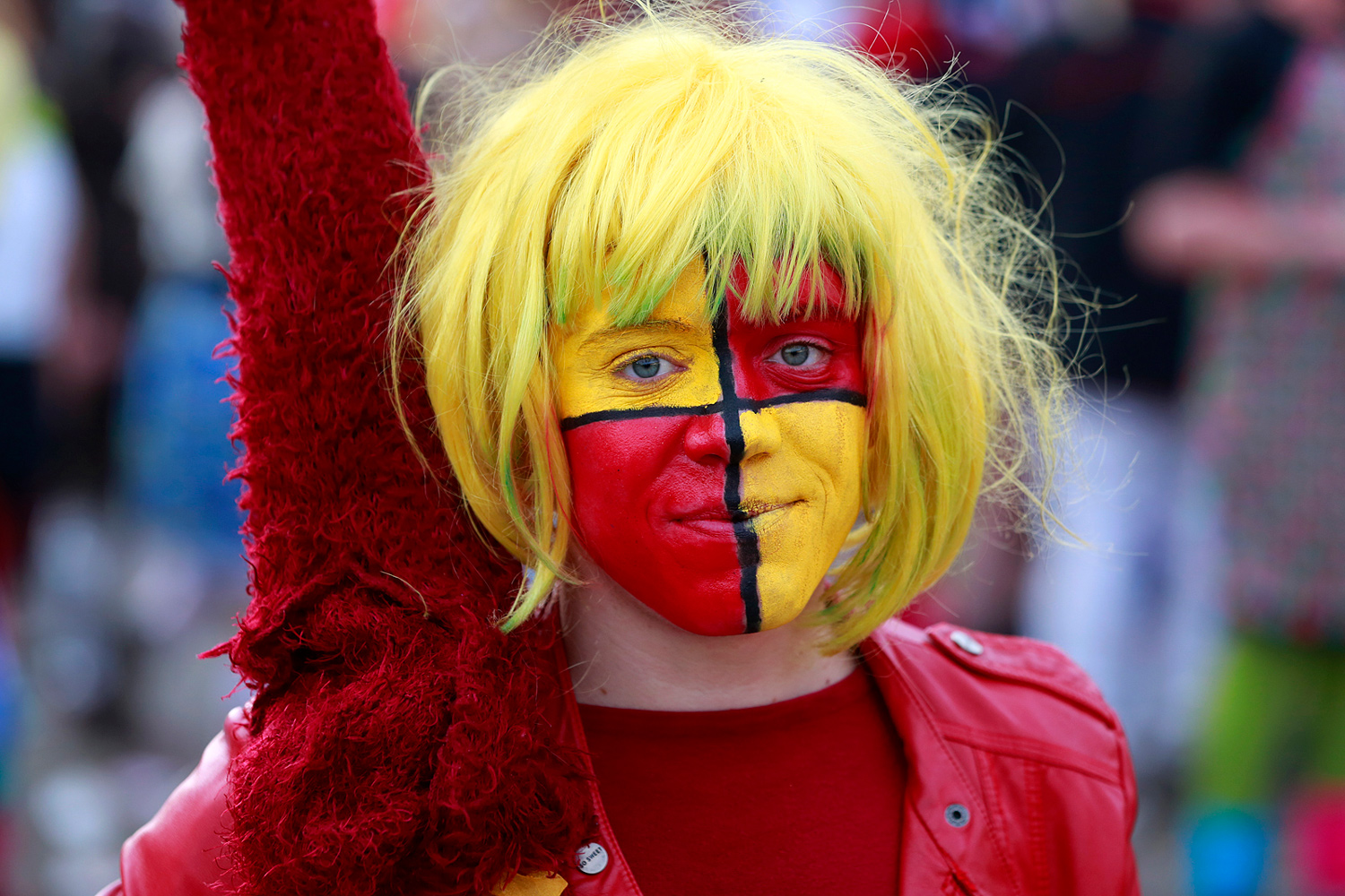 Portrait d'une jeune carnavalière levant le bras, portant une perruque blonde et un maquillage partagé en rouge, jaune, noir et bleu aux couleurs du drapeau belge, lors de la Bande de Pitgam du Carnaval de Dunkerque.