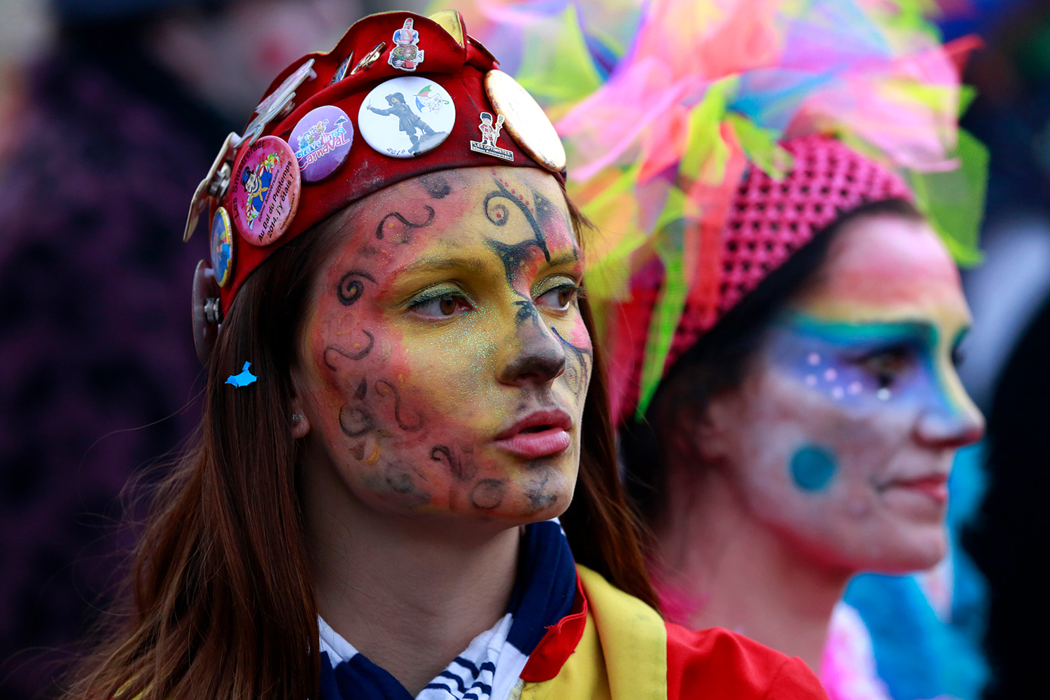 Portrait d'une carnavalière au visage peint de motifs jaunes, rouges et noirs, coiffée d'un casque rouge décoré de nombreux badges.