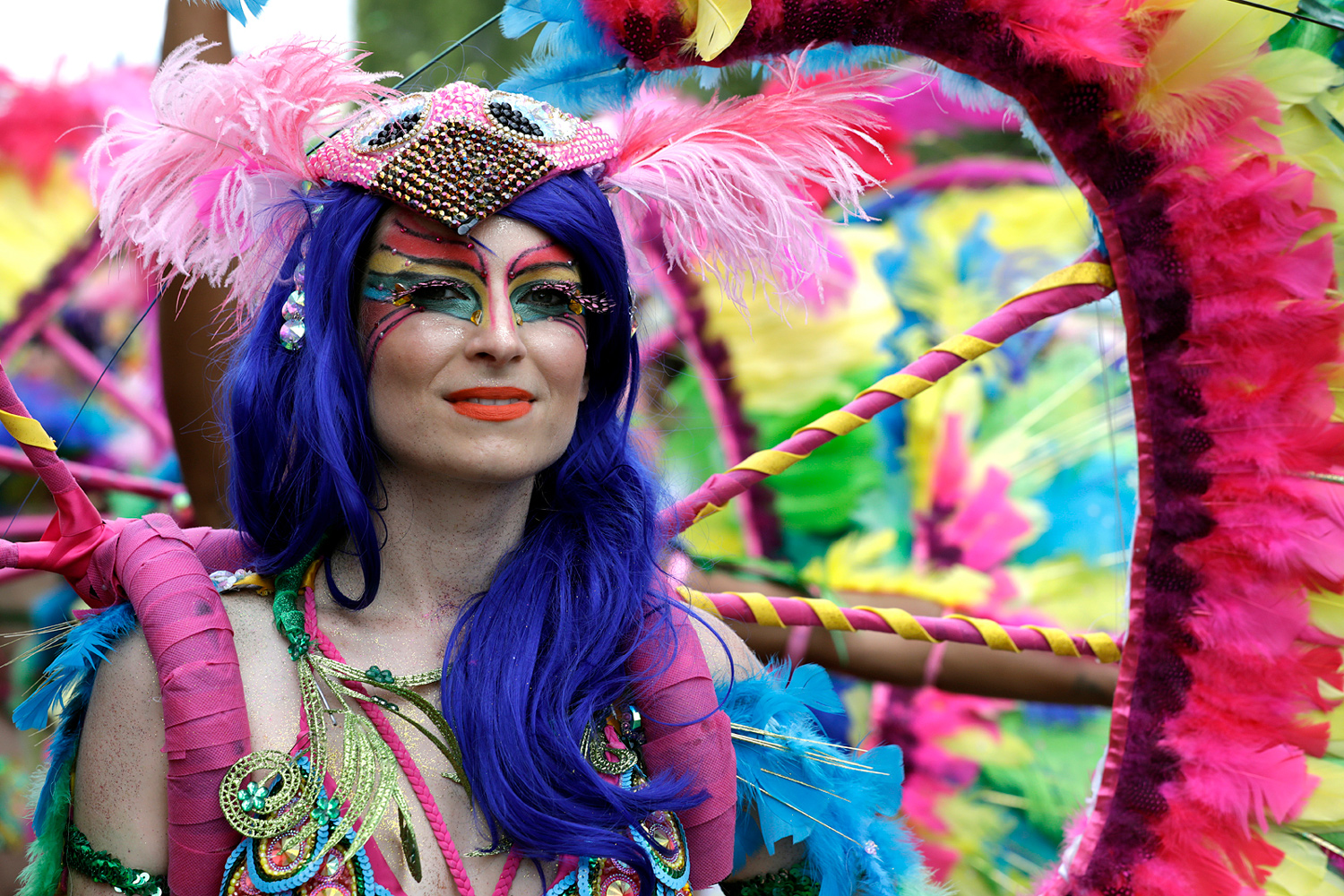 Portrait d'une danseuse portant une longue perruque bleue et un costume orné de plumes roses et bleues.
