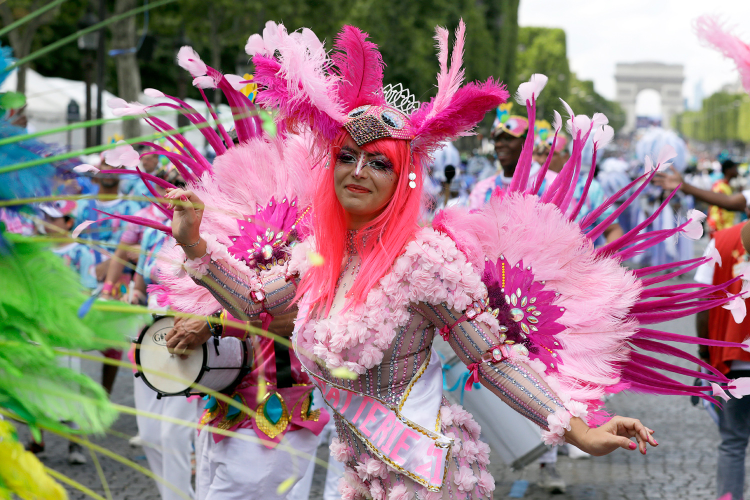Danseuse en costume rose à plumes avançant dans le défilé avec les Champs-Élysées en arrière-plan.