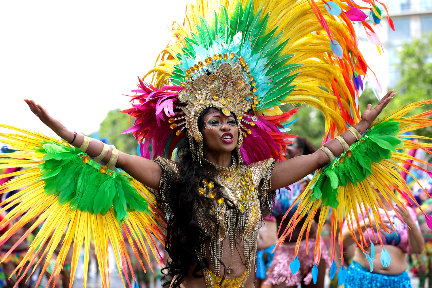 Portrait d'une danseuse portant une immense coiffe jaune, verte et rose ornée de plumes, les bras ouverts pendant le défilé.