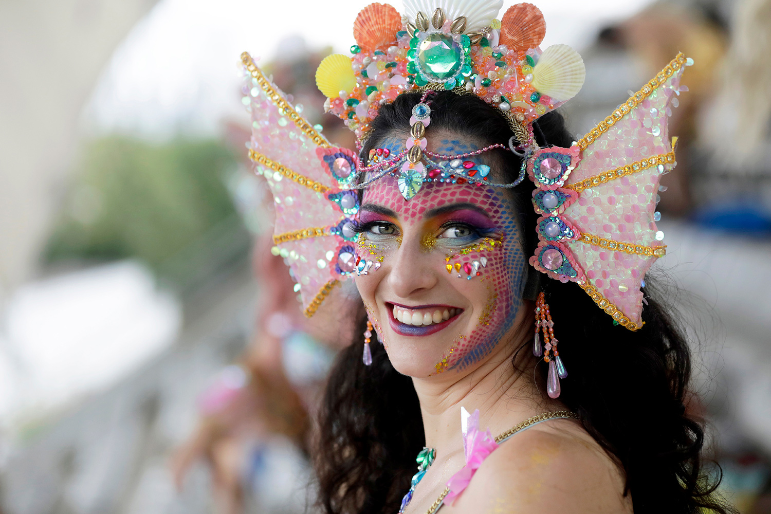 Portrait d'une danseuse souriante portant une coiffe rose ornée de coquillages et un maquillage aux couleurs pastel.