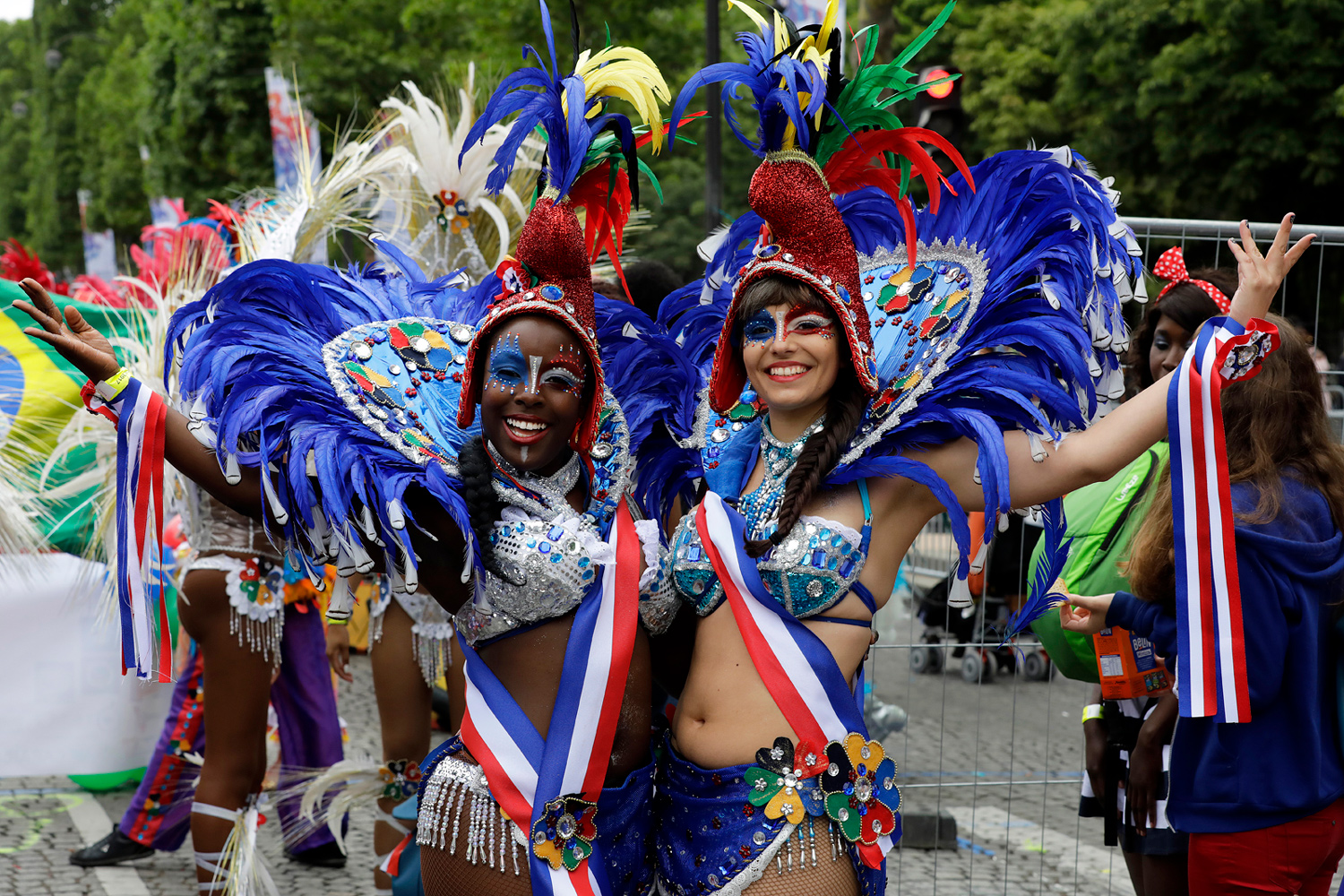 Deux danseuses souriantes posent côte à côte dans des costumes bleus à plumes pendant le défilé.