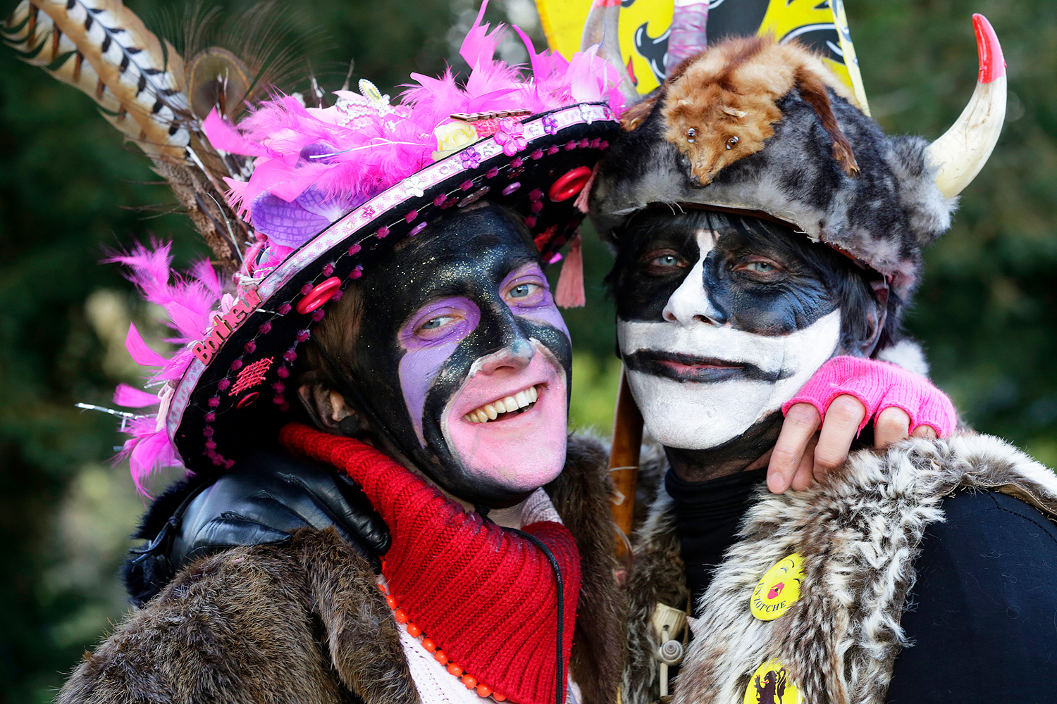 Deux carnavaleux souriants, maquillés de noir et de blanc, posent enlacés, coiffés d'un chapeau fleuri et d'un casque à cornes.