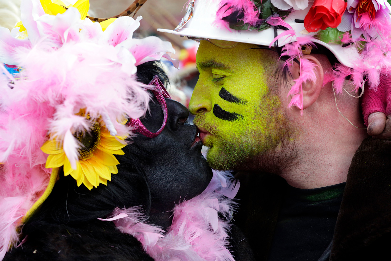 Deux carnavaleux s'embrassent, l'un maquillé en noir et l'autre en vert, coiffés de chapeaux décorés de plumes et de fleurs.